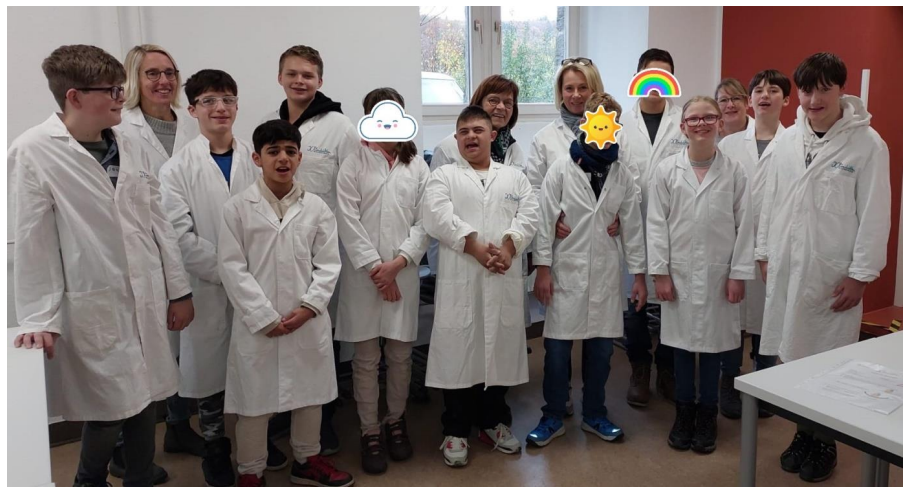 Auf dem Bild sieht man eine Schulklasse mit ihren Lehrerinnen in Laborkitteln.