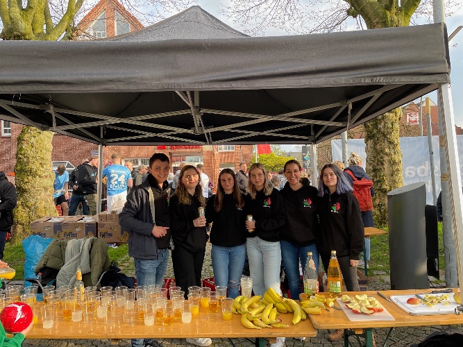 Auf dem Bild sind Mitarbeiterinnen und Mitarbeiter der Schülerfirma Break n´Bite hinter der Versorgungsstand. Auf dem Tisch des Standes stehen Getränke, Bananen und Äpfel für die Läufer bereit.
