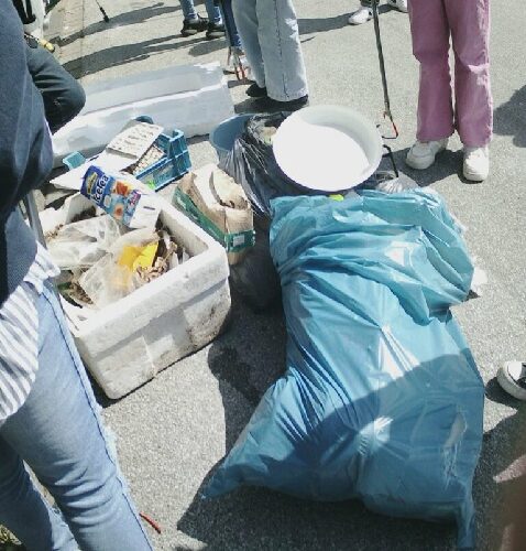 Eine große blaue Mülltüte sowie mehrere Behälter voller gesammeltem Müll stehen auf dem Boden vor den Füßen zweier Schüler, die eine Greißzange in der Hand halten. 