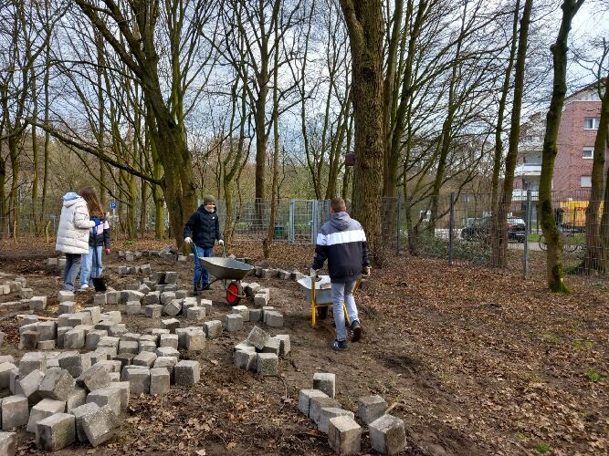 Zwei Jungen der Schulgarten-AG der Jahrgangsstufe 6 und zudem einige Mädchen der Schulgarten-AG der Jahrgangsstufe 7 arbeiten in der Freifläche des Barfußpfads umgeben von zahlreichen Pflastersteinen, die sie bereits dorthin transportiert haben.