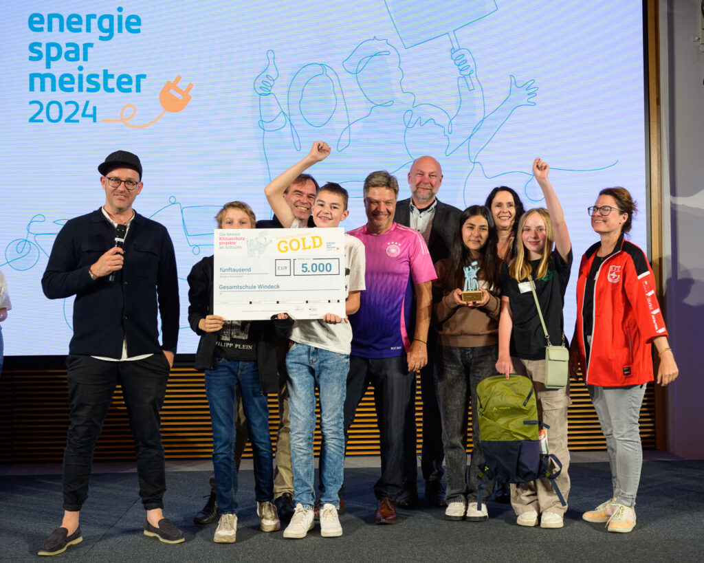 Gruppenfoto, auf dem Schüler:innen der Gesamtschule Windeck einen Scheck über 5000€ in die Höhe halten. Ebenfalls zu sehen sind Bundeswirtschaftsminister Robert Habeck und die Moderator:innen der Auszeichnungsfeier.