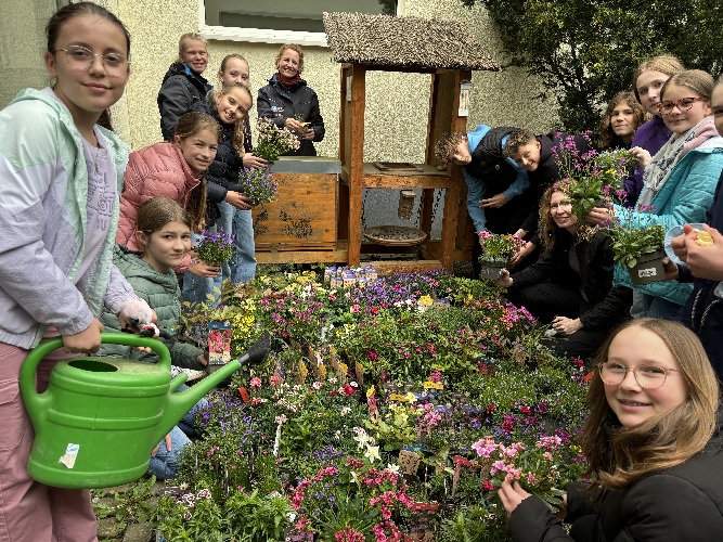 Das Bild zeigt Schülerinnen und Schüler inmitten von den zu pflanzenden Blumen in einem Außenbereich der Schule.