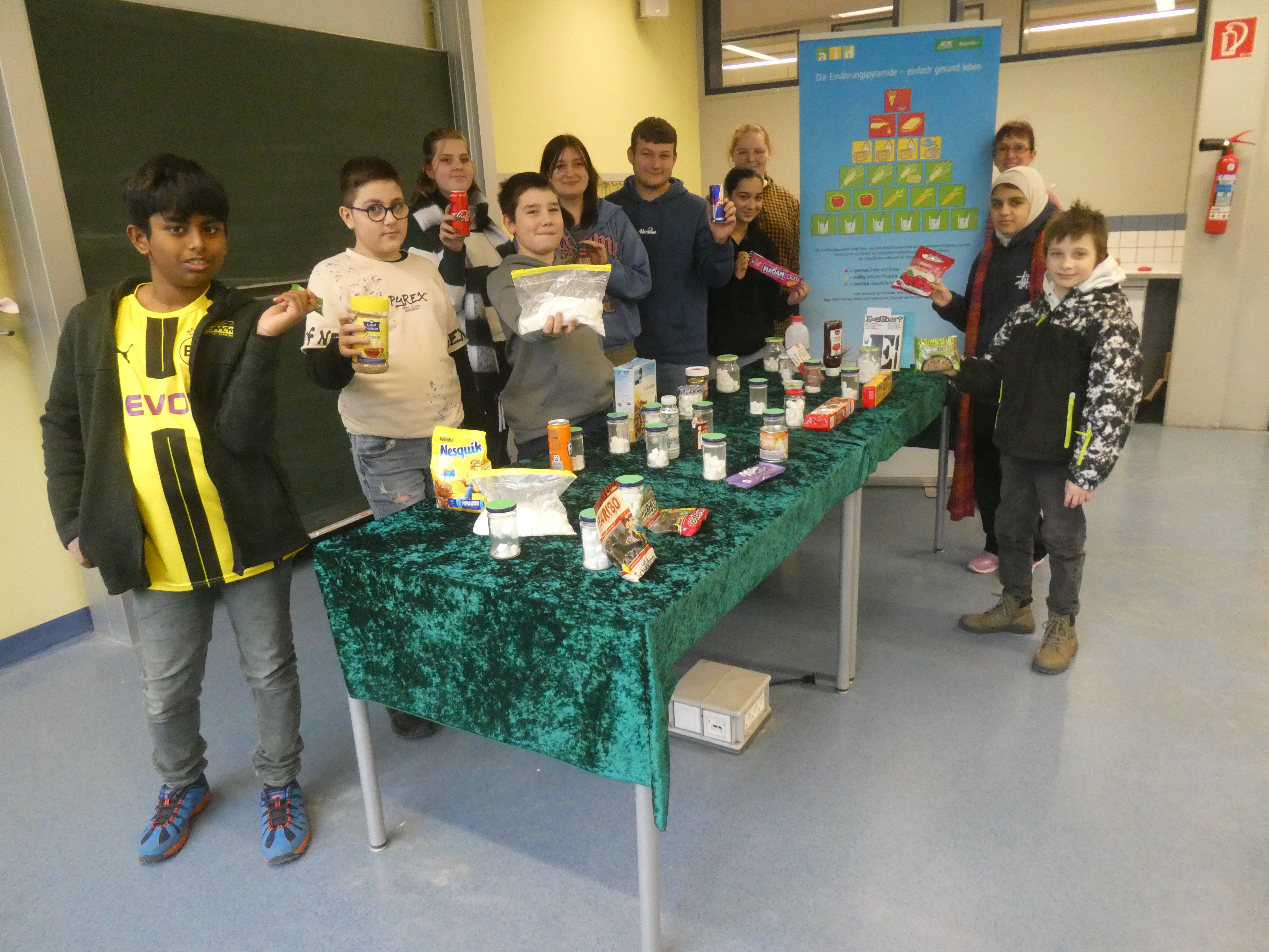 Das Bild zeigt die Fairness-Detektive und Kiosk-Helfer*innen an einem Tisch mit Lebensmitteln und Würfelzuckerportionen, hinten steht Frau Schieffers vor der Ernährungstabelle.