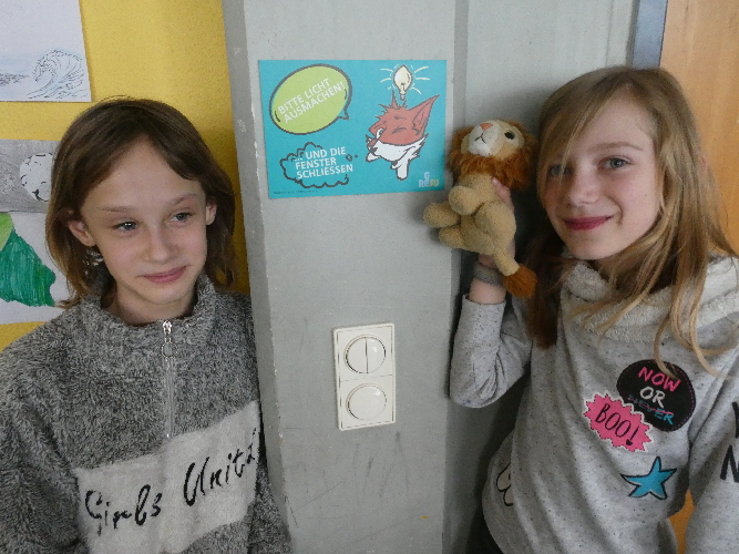 Das Bild zeigt zwei Schülerinnen rechts und links neben dem Schild mit dem Klima-Fuchs, das im Klassenraum direkt an den Lichtschaltern angebracht ist.