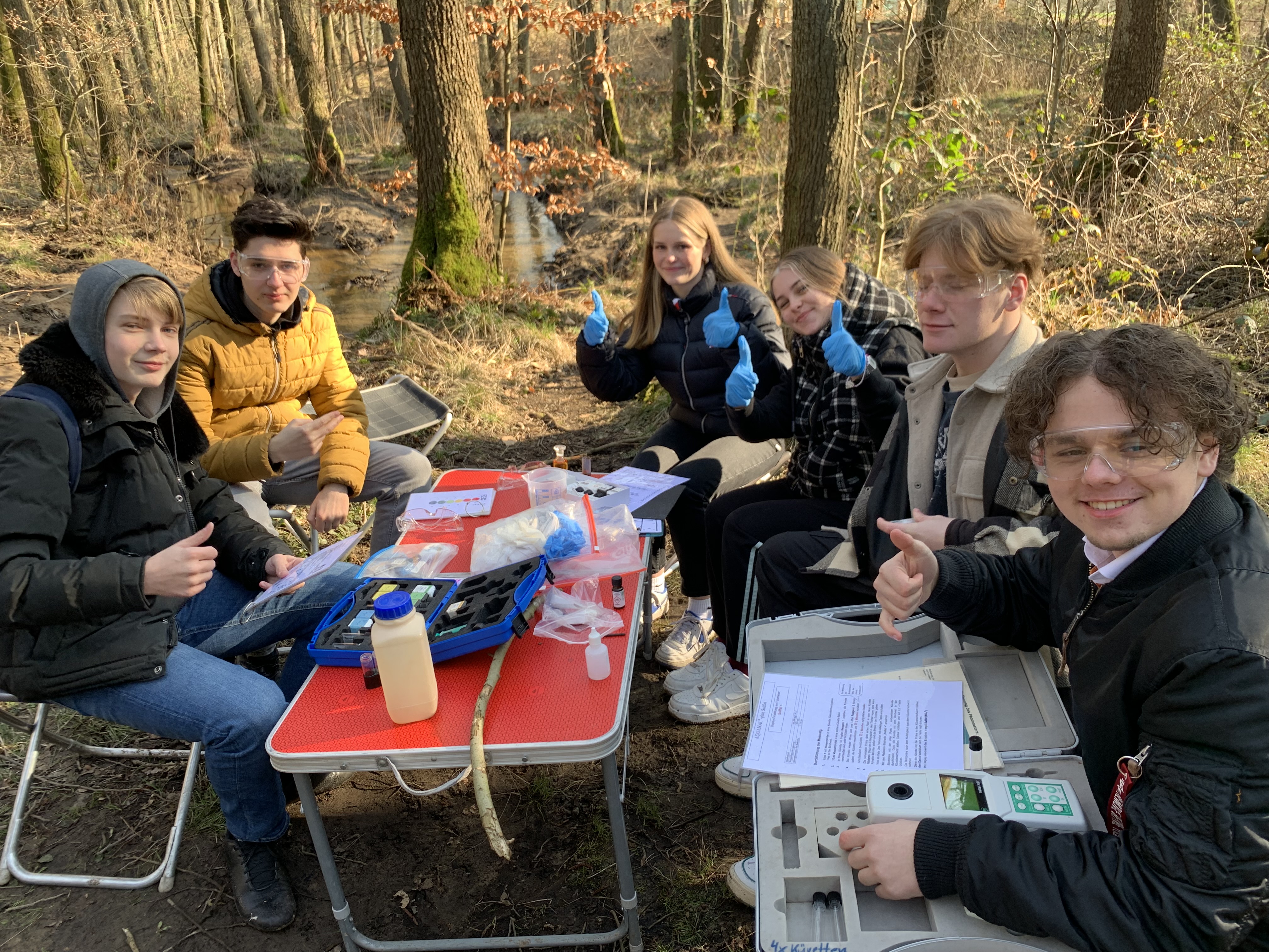 Das Bild zeigt sechs Schüler*innen, die im Wald direkt neben dem Bach an einen Tisch sitzen, an dem Sie mit verschiedenen Geräten Wasseranalysen durchführen.