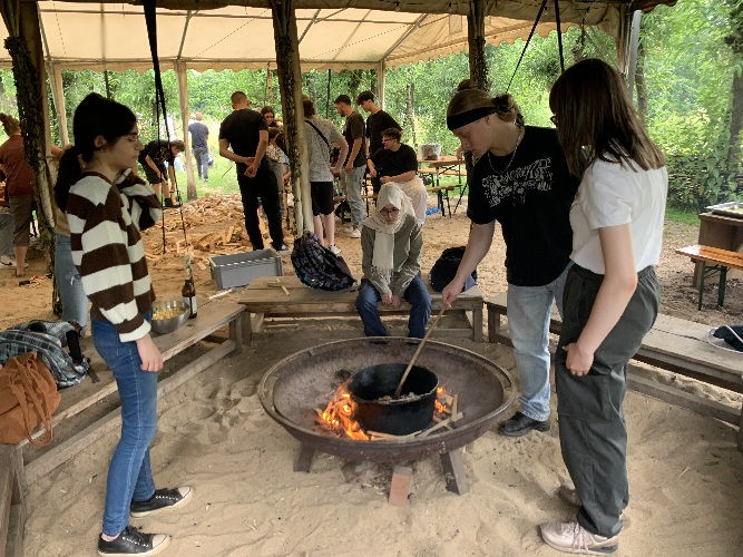 Im Vordergrund stehen drei Schüler*innen am offenen Feuer und kochen Gemüsesuppe, im Hintergrund hackt eine größere Schüler*innen-Gruppe Feuer-Holz.