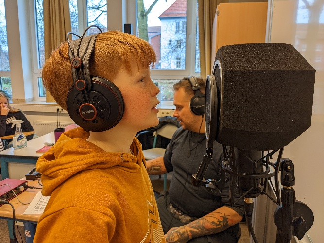 Ein rothaariger Junge mit Kurzhaarschnitt und schwarzen Kopfhörern steht rappend vor einem schwarzen Mikrophon.