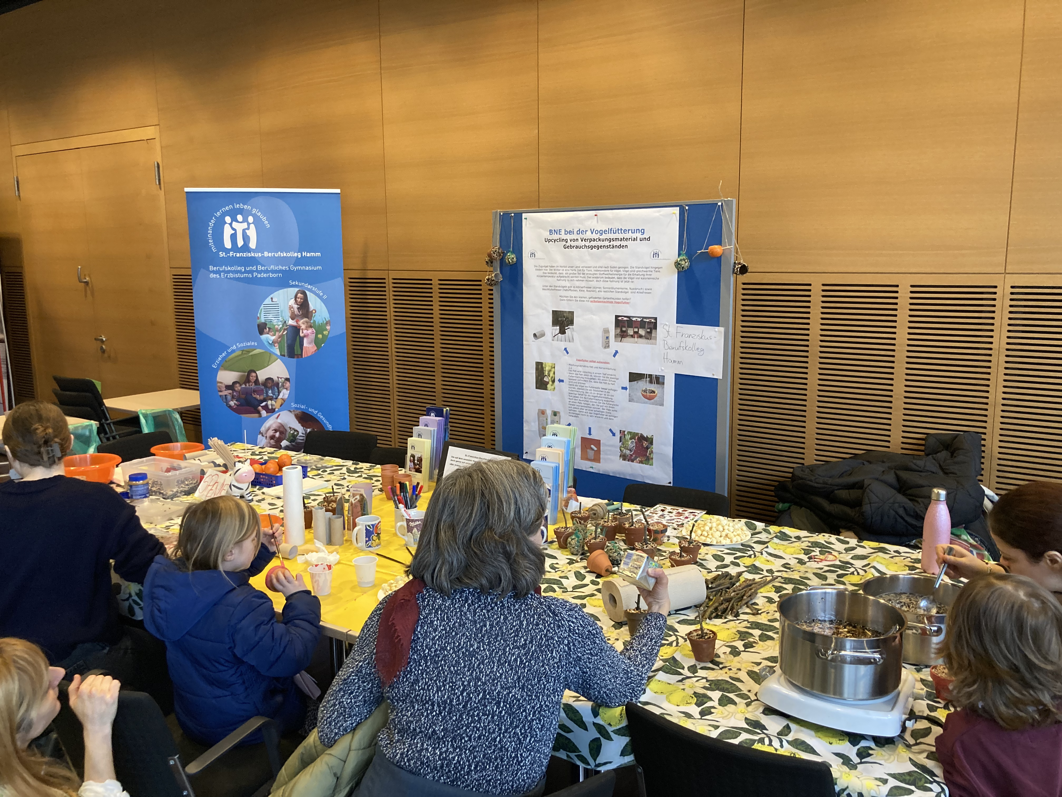 Zu sehen ist der Stand des SFBK bei der 2. Nachhaltigkeits- und Mitmachmesse in Hamm. Um den Tisch, auf dem sich verschiedene Materialien befinden, sammeln sich Personen, die Vogelfutter herstellen. Im Hintergrund ist eine Pinnwand mit wichtigen Informationen zu BNE bei der Vogelfütterung zu erkennen.