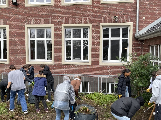 Vor dem Schulgebäude sieht man mehrere SchulerInnen, die das Beet von Brennnesseln und Grasern befreien.