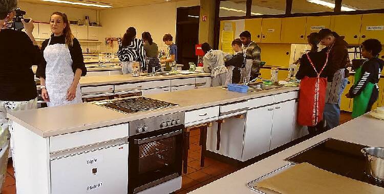 Die Lehrerin beantwortet in der Lehrküche der Schule Fragen vor laufender Kamera. Im Hintergrund arbeiten Schüler und Schülerinnen zwischen den Küchenzeilen an der Zubereitung ihrer Rezepte. 