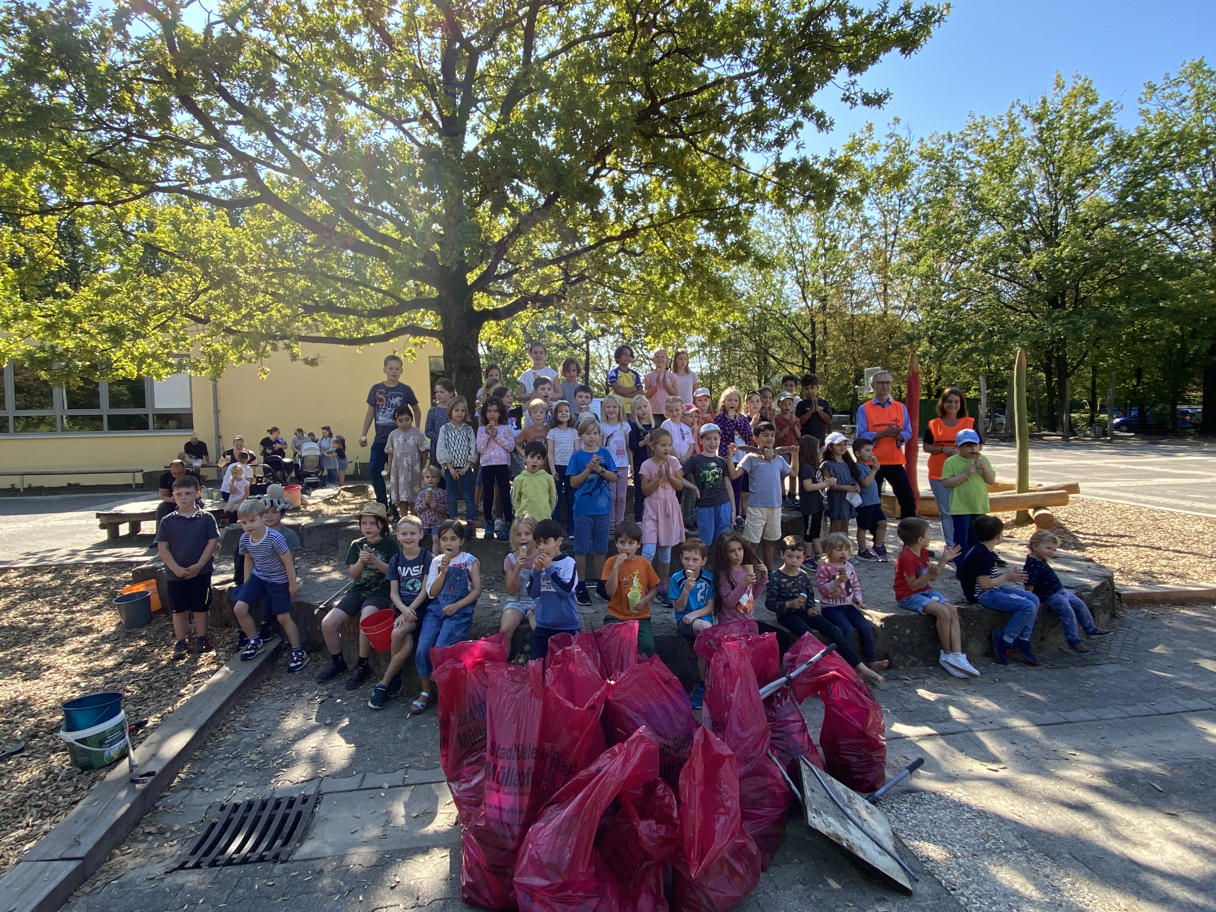 Auf dem Foto sind viele Kinder unter einer Eiche zu erkennen, die ein Eis in der Hand haben. Rechts im Hintergrund sieht man die Lehrerin mit dem Bezirksbürgermeister in orangen Stiftsschulleibchen. Im Vordergrund sind viele rote Mülltüten vollgepackt mit Müll.