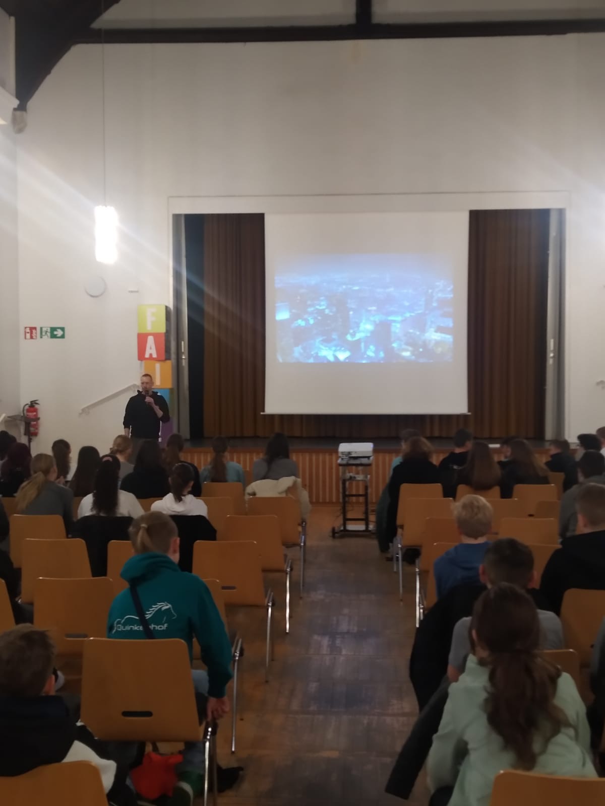 Man sieht die Rücken der Schüler/innen, eine Leinwand und den Redner mit Mikrofon in der Hand.