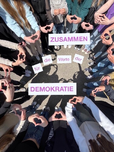 In der Mitte steht Zusammen für die Werte der Demokratie. Drumherum stehen Schüler im Kreis, die ihre Hände zu Herzen formen.