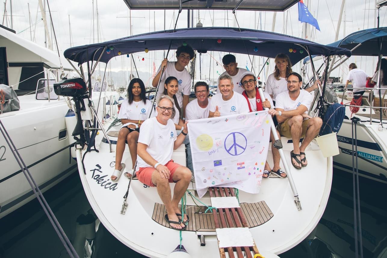 Das Foto zeigt Schüler:innen des Schillergymnasiums, Lehrkräfte und Crew-Mitglieder, die auf dem Heck eines Segelschiffes sitzen und eine selbstgestaltete PEACE-Flagge ins Bild halten.